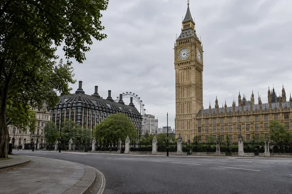 A quiet overcast street view of Elizabeth Tower and the Palace of Westminster, with dark leafy branches framing the left side, a curved pavement kerb and broad…