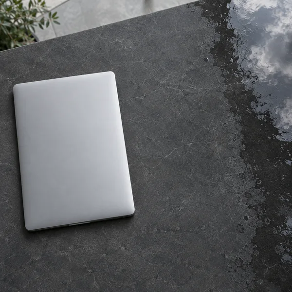 A closed silver laptop rests diagonally on a charcoal stone terrace counter, with a rain-darkened reflective strip along the right edge and leafy branches…