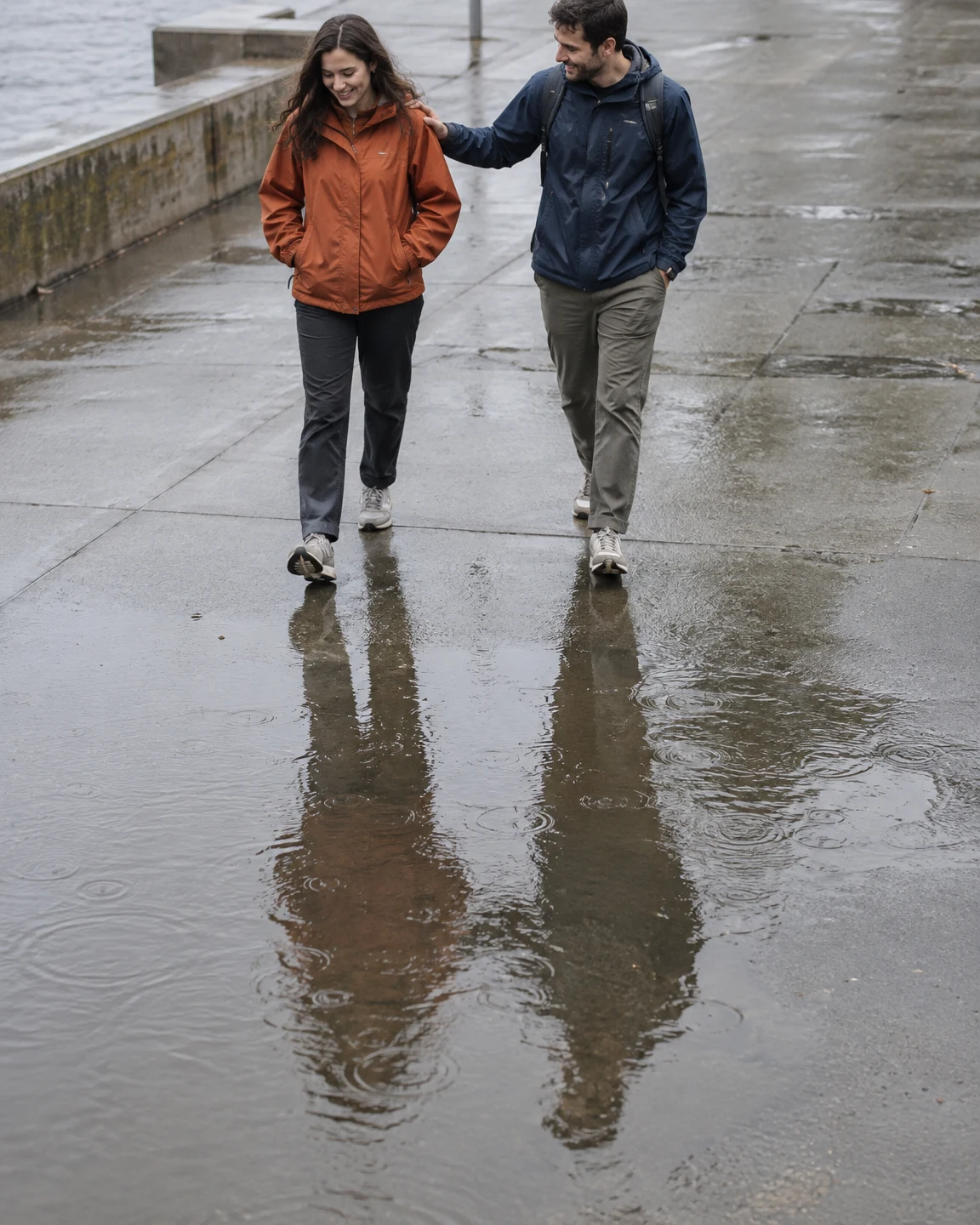 A woman in a burnt-orange jacket and a man in a navy jacket walk across wet concrete, with the man resting a hand on her shoulder.