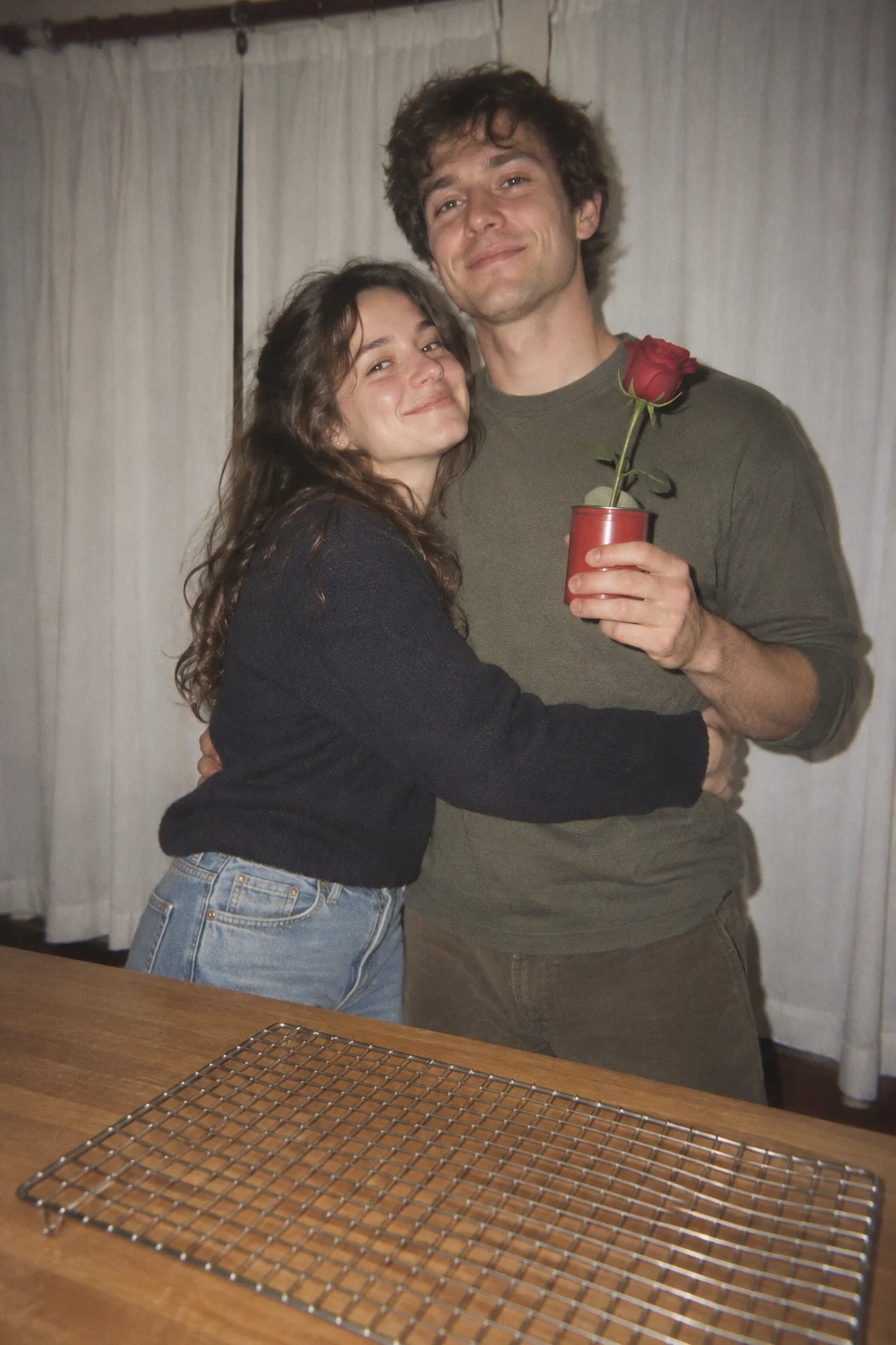 A smiling young couple embraces in front of white curtains, with the man holding a red rose in a small red can; a large metal cooling rack lies diagonally…