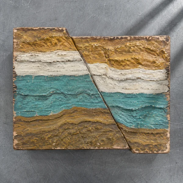 An overhead view of a worn carved-wood geological relief on a scratched grey metal workbench, with turquoise, chalk-white and ochre layers offset along a…