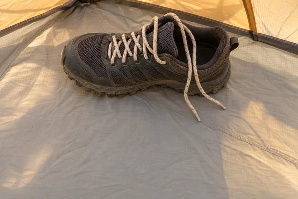 A single charcoal trail shoe with cream laces rests on a pale gray ripstop tent floor beneath warm amber tent panels; the toe points left and the loose lace…