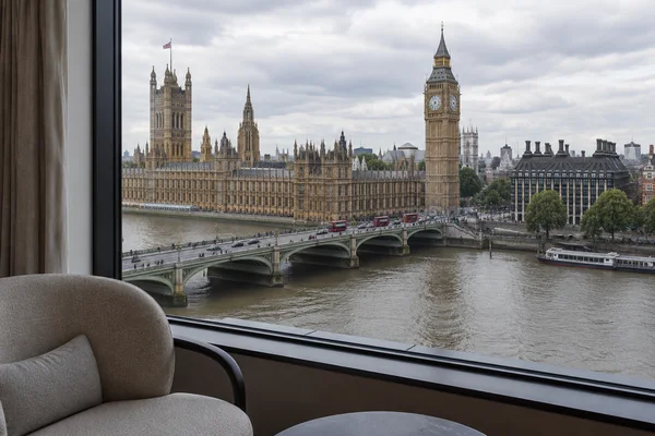 A view across the Thames toward Elizabeth Tower and the Palace of Westminster, with Westminster Bridge crossing the foreground; a pale rounded lounge chair and…