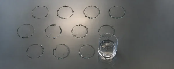 An overhead view of a smoke-grey glass tabletop shows rows of damp circular marks and a clear low tumbler with a little water remaining, lit from beneath.