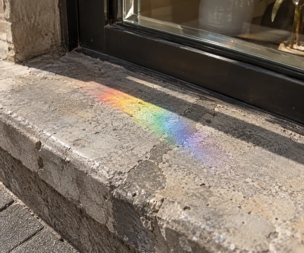 A diagonal band of red, orange, yellow, green, blue and violet light falls across a worn stone threshold beneath a dark metal-framed window, crossing a grey…