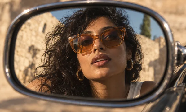 A woman with tousled dark curls, amber sunglasses and gold hoop earrings appears in a close reflection in a dark chrome car side mirror, with a sunlit stone…