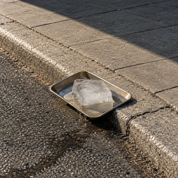 A cloudy, partly melted block of ice sits in a shallow metal tray balanced across a concrete curb, with pooled water spilling onto the dark asphalt in warm…