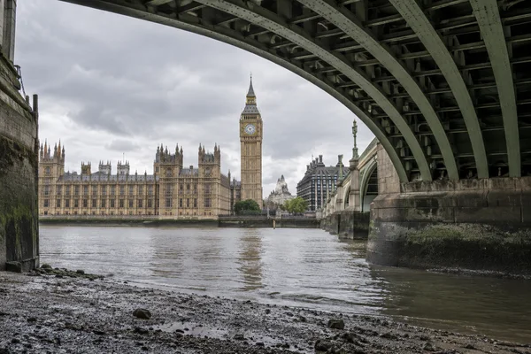 A low viewpoint from a damp, gravelly Thames foreshore looks beneath the green iron structure of Westminster Bridge toward Elizabeth Tower and the Palace of…