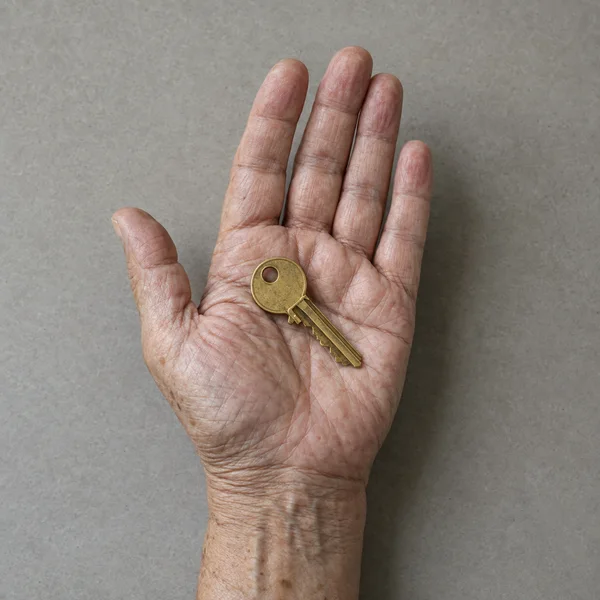 An open, wrinkled hand holds a worn brass key diagonally across the palm against a plain gray surface.