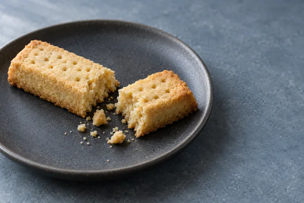 Two uneven pieces of golden shortbread with visible broken crumb and loose crumbs sit on a dark ceramic plate against a blue-grey tabletop, with open space to…