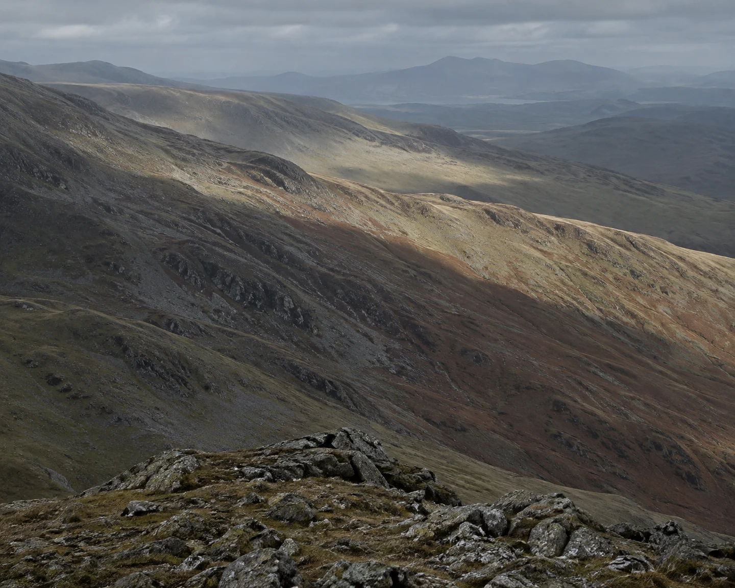 A rocky foreground ridge overlooks layered upland slopes of grey scree, olive grass and rust-coloured bracken.