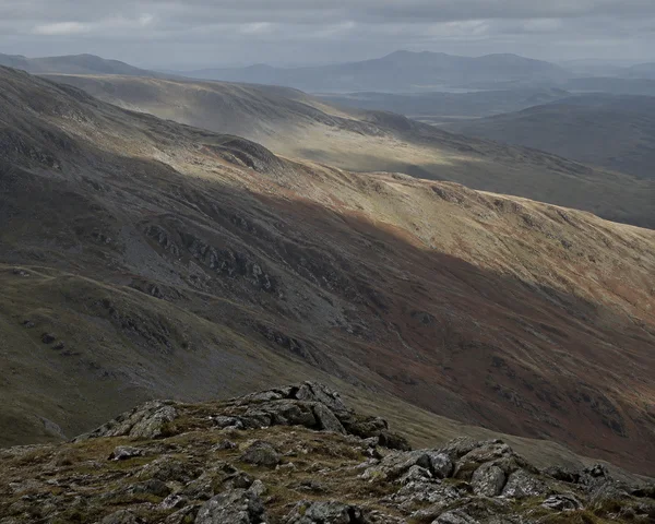 A rocky foreground ridge overlooks layered upland slopes of grey scree, olive grass and rust-coloured bracken.