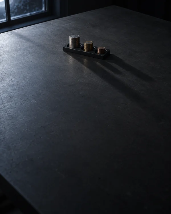 A narrow black tray holding three unequal stacks of silver- and brass-toned coins sits near the upper center of a dark charcoal table, with long shadows…