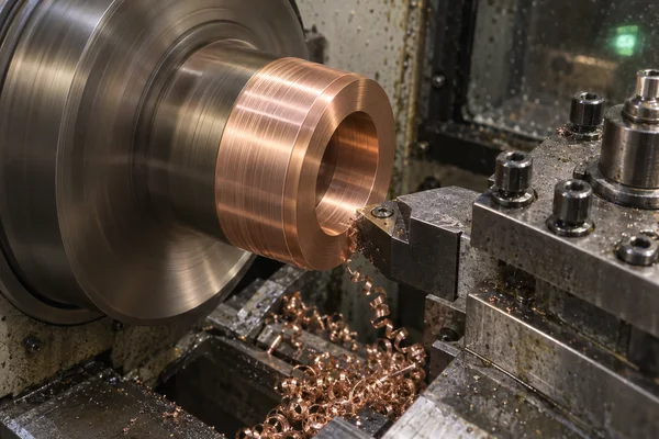 A copper collar rotates on a lathe as a cutting insert contacts its rim, with curled copper swarf collected beneath the tool and a small green indicator light…
