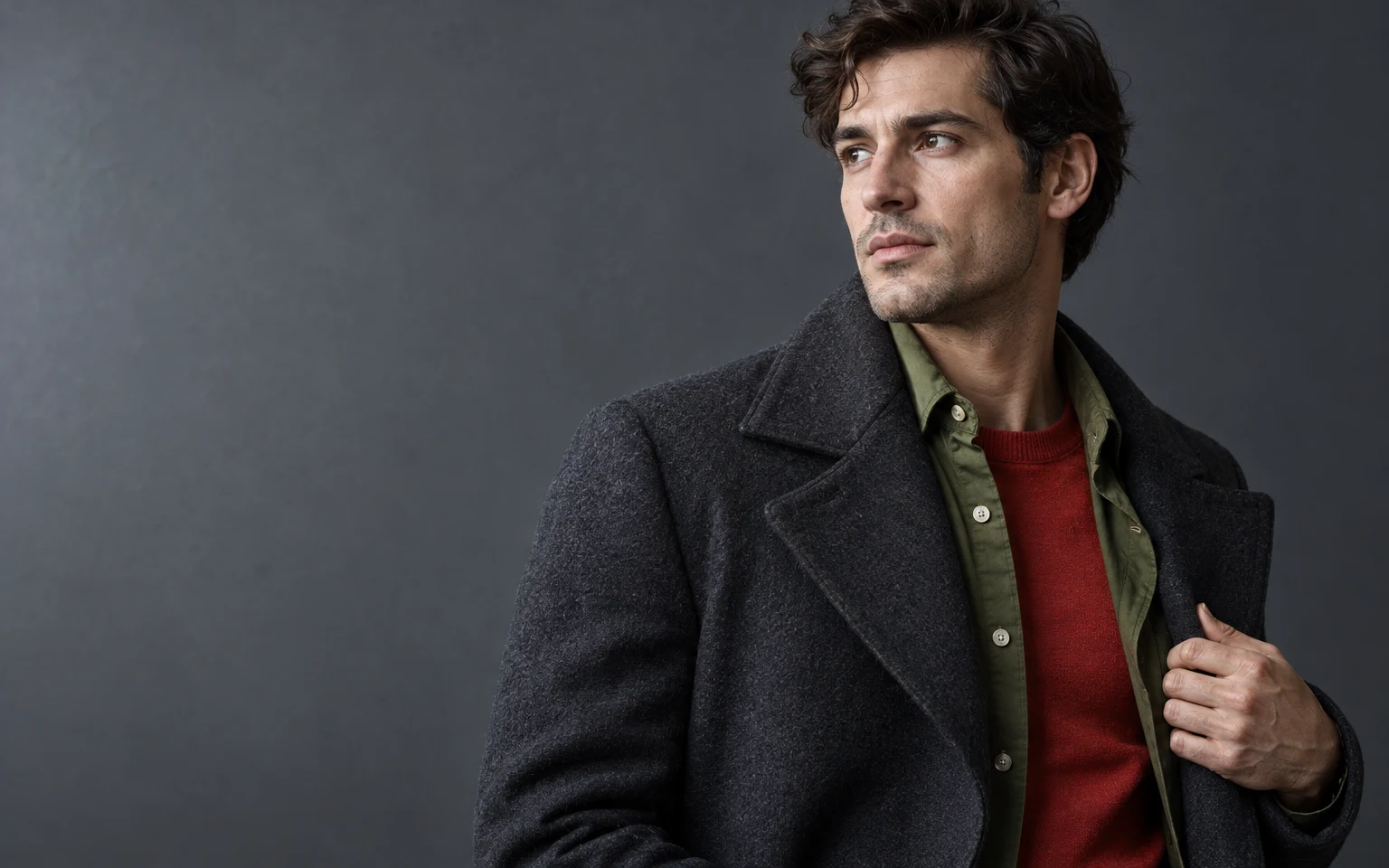 A dark-haired adult in a charcoal coat over an olive-green shirt and red crewneck sweater stands against a slate-grey backdrop, holding the coat lapel with one…