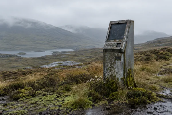 A weathered grey metal terminal kiosk with a dark, dusty screen and keypad stands among moss, tussock grass and small white flowers on a damp moorland slope…