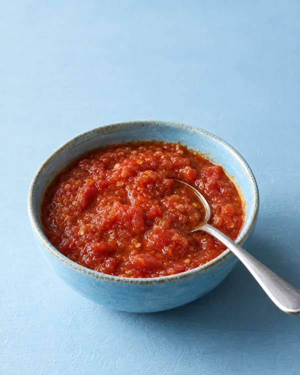 A speckled sky-blue ceramic bowl filled with chunky red tomato sauce sits on a pale blue tabletop, with a stainless-steel spoon partly submerged and angled…