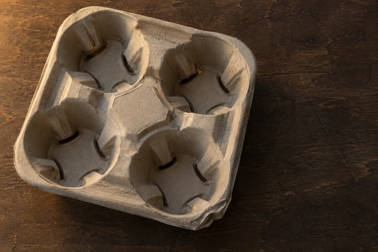 A grey-beige molded-pulp four-cup drinks carrier rests diagonally on a dark brown plywood worktable, lit by warm directional light with open tabletop to the…