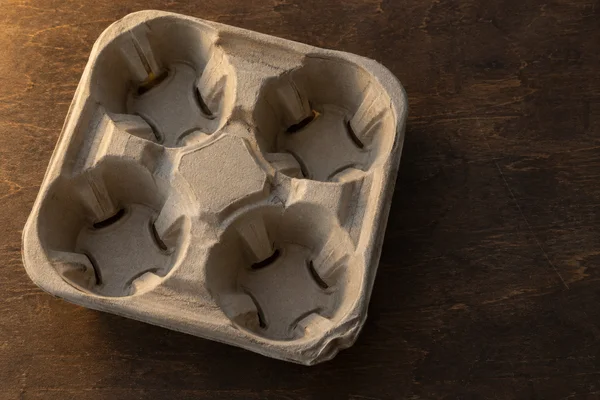 A grey-beige molded-pulp four-cup drinks carrier rests diagonally on a dark brown plywood worktable, lit by warm directional light with open tabletop to the…