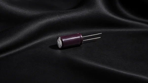 A dark plum cylindrical capacitor with a silver scored end cap and two straight metal leads lies on black satin, with broad folds and a crisp shadow.