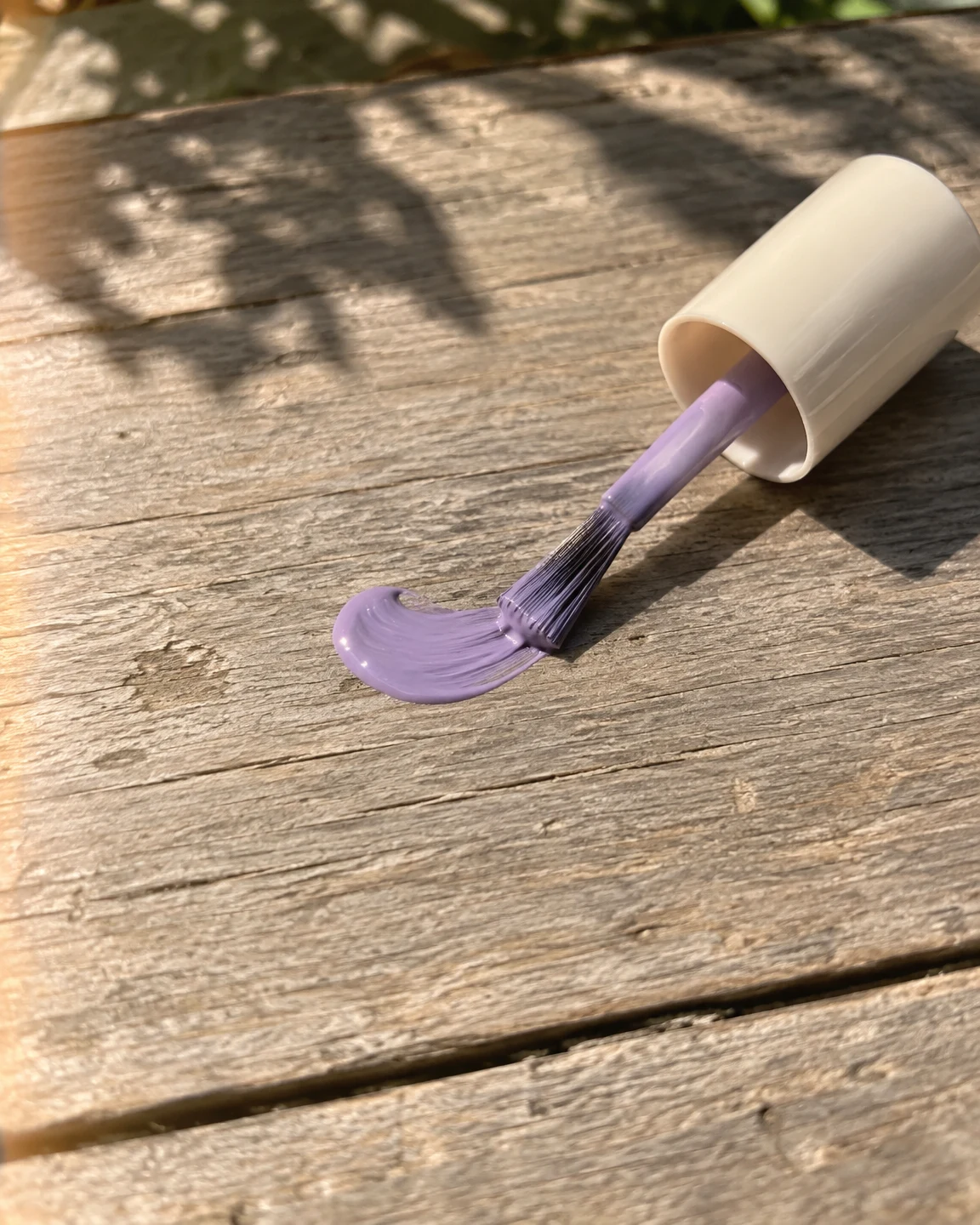 A nail-polish brush with a plain ivory cap rests on its side on a weathered wooden table, its lilac-coated bristles touching a glossy crescent of polish in…