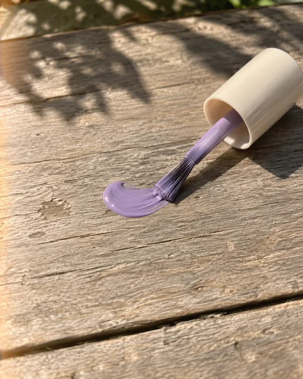 A nail-polish brush with a plain ivory cap rests on its side on a weathered wooden table, its lilac-coated bristles touching a glossy crescent of polish in…
