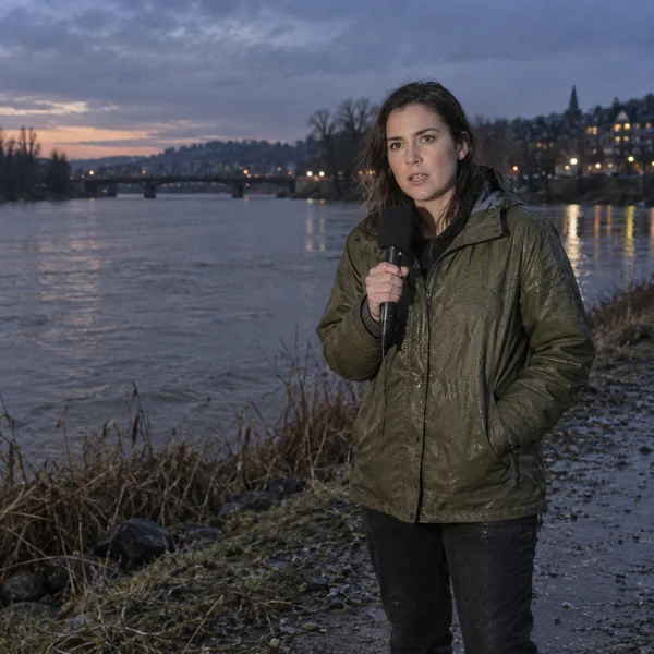 A woman speaks into a handheld microphone on a damp riverside embankment at dusk, with the river and distant illuminated buildings behind her.