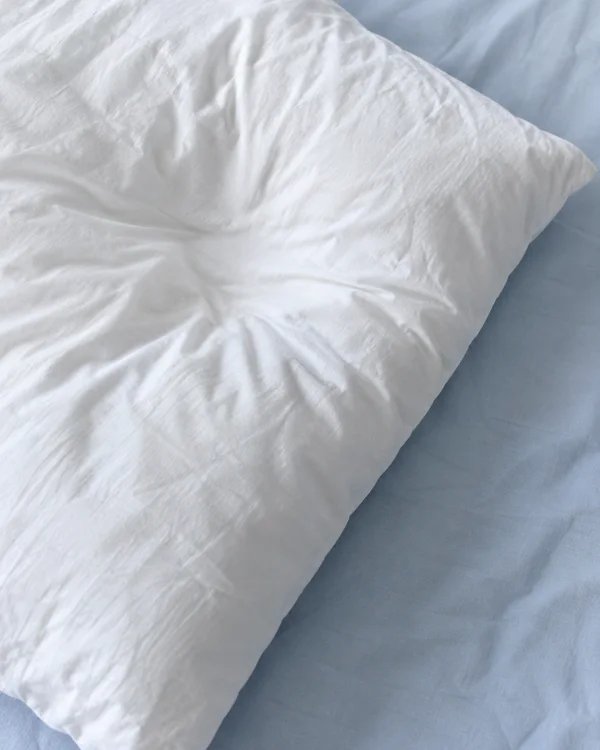 Close-up of a white cotton pillow with radiating creases, lying diagonally on a pale blue sheet.