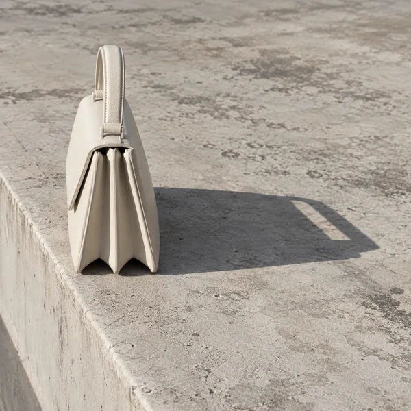 An ivory leather handbag stands end-on on a weathered pale concrete parapet, with its upright handle and folded gussets casting a long angular shadow across…