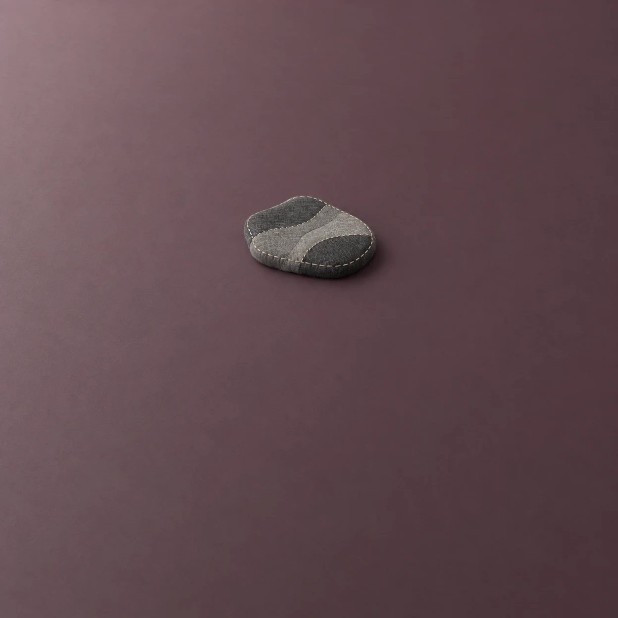 A small charcoal-and-gray stitched textile form with an irregular rounded outline rests on a muted plum surface, casting a soft shadow.