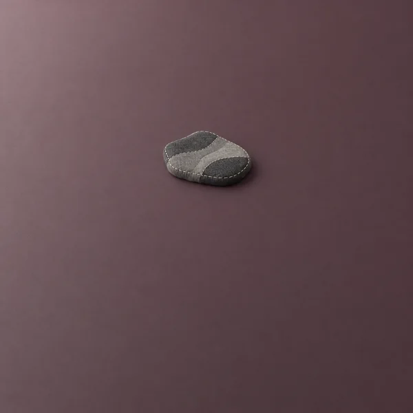 A small charcoal-and-gray stitched textile form with an irregular rounded outline rests on a muted plum surface, casting a soft shadow.