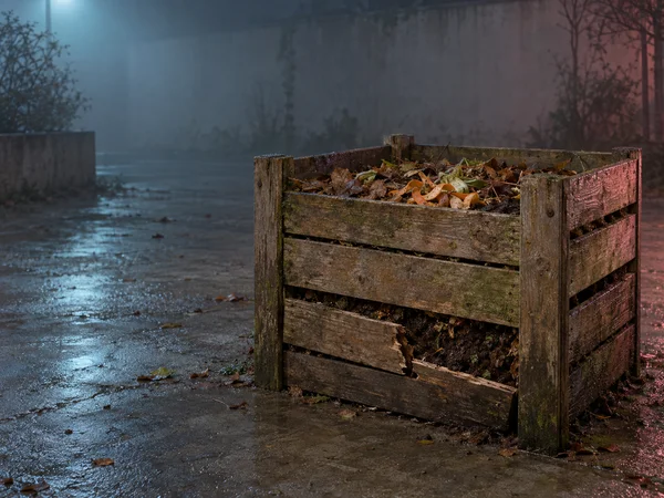 A weathered wooden compost bin filled with damp leaves and vegetable scraps sits on wet concrete at night, with a broken lower slat exposing dark compost.