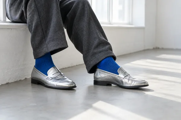 An adult sits on a white-painted window ledge, shown from the knees down in charcoal trousers, vivid cobalt ribbed socks and silver loafers; one shoe is…