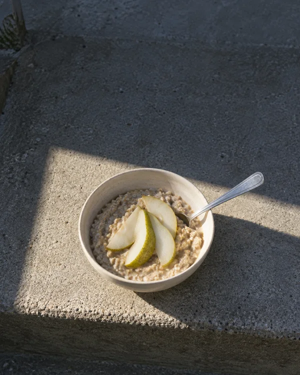 A cream bowl of oatmeal topped with three pear slices sits on a weathered concrete garden step in a sharply edged patch of sunlight, with a teaspoon resting…