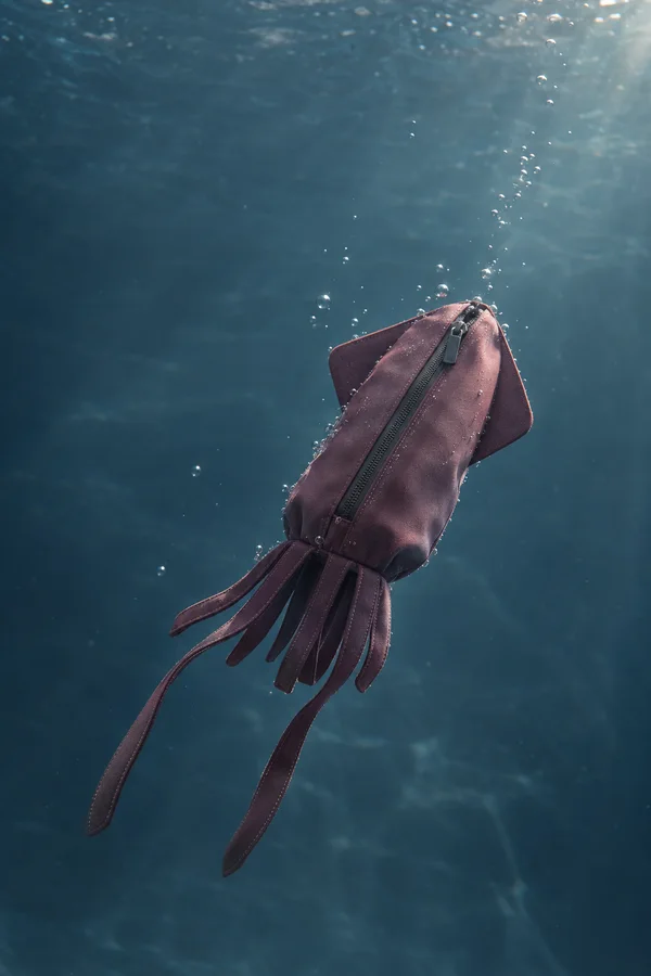 A plum squid-shaped waterproof pouch floats diagonally underwater, with a long front zipper, angular side fins, short fabric arms and two longer trailing…