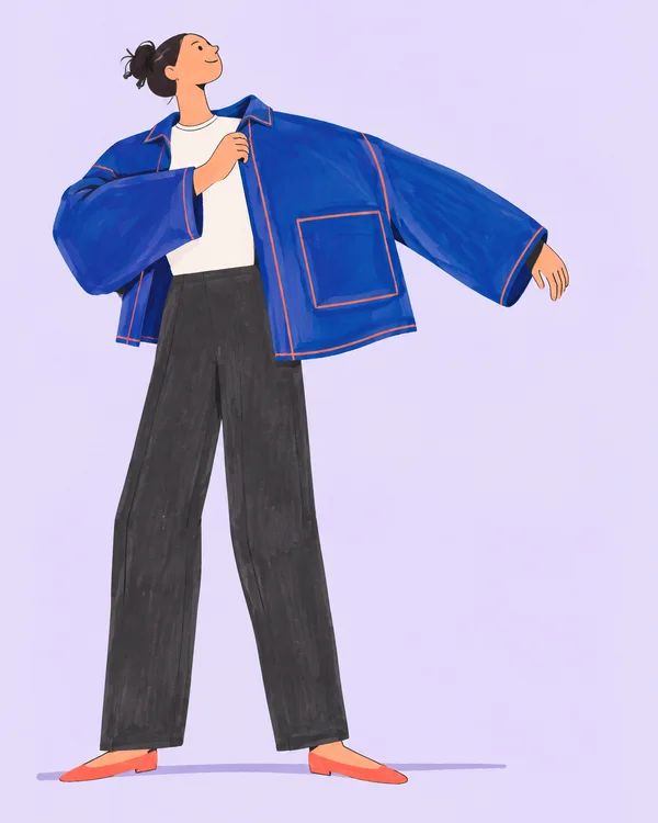 A smiling garment maker stands with chin lifted, wearing an oversized cobalt-blue jacket with coral seam lines and a large patch pocket, charcoal trousers and…