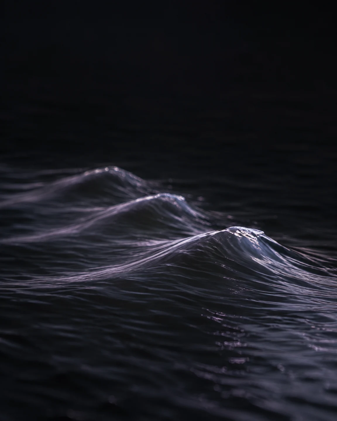 A dark, close view of several lavender-lit wave crests crossing rippled black water, with a large area of nearly black water above them.