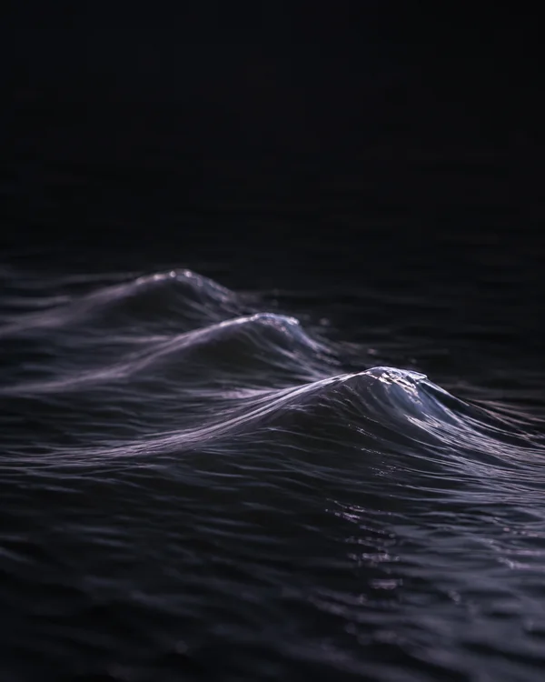 A dark, close view of several lavender-lit wave crests crossing rippled black water, with a large area of nearly black water above them.