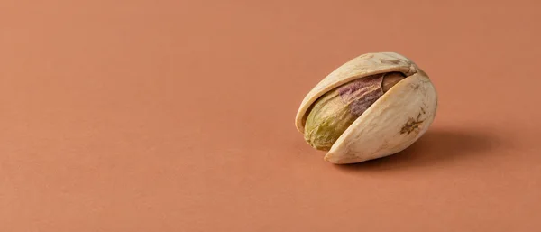 A naturally split pistachio with an olive-green kernel and muted purple skin edge rests on a matte pale terracotta surface, positioned just right of center…
