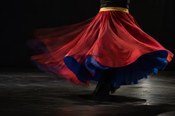 A dancer’s red flared skirt with cobalt-blue lining and a yellow waistband sweeps across a dark rehearsal stage, with several translucent hem positions visible…