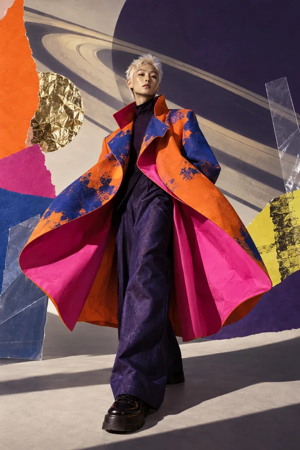 A short-haired model in an orange-and-cobalt coat with vivid fuchsia lining stands against a studio backdrop crossed by broad planetary-ring shapes.