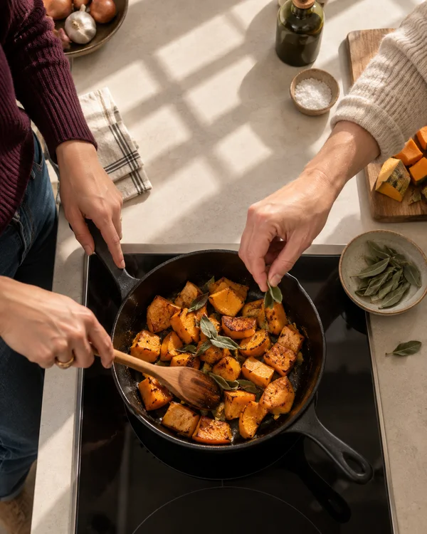 An overhead kitchen scene shows golden squash pieces and sage in a black skillet on an induction hob.