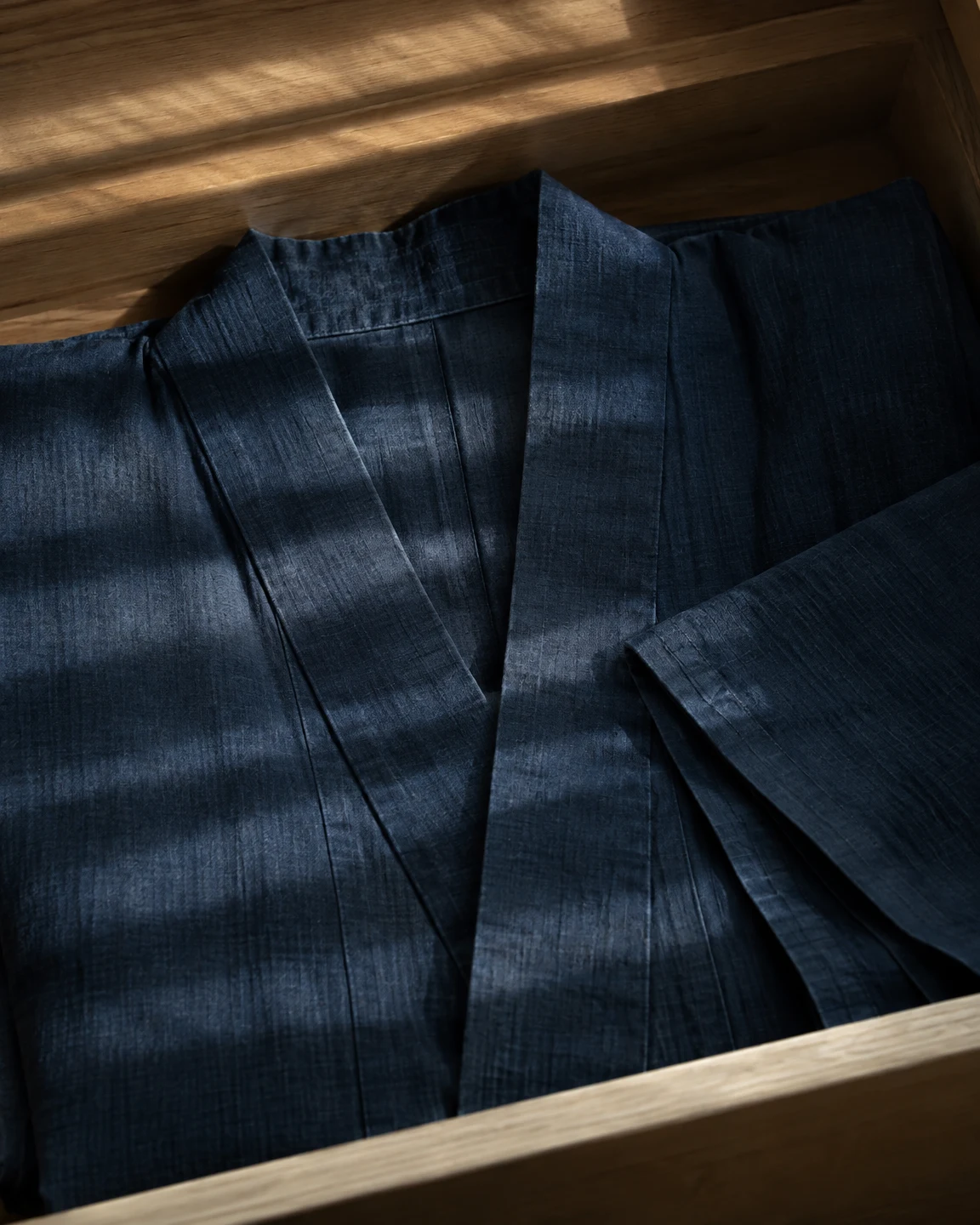 A dark indigo kimono lies inside a wooden drawer, its overlapping collar and folded sleeve visible beneath bands of light and shade; warm wood frames the cloth…