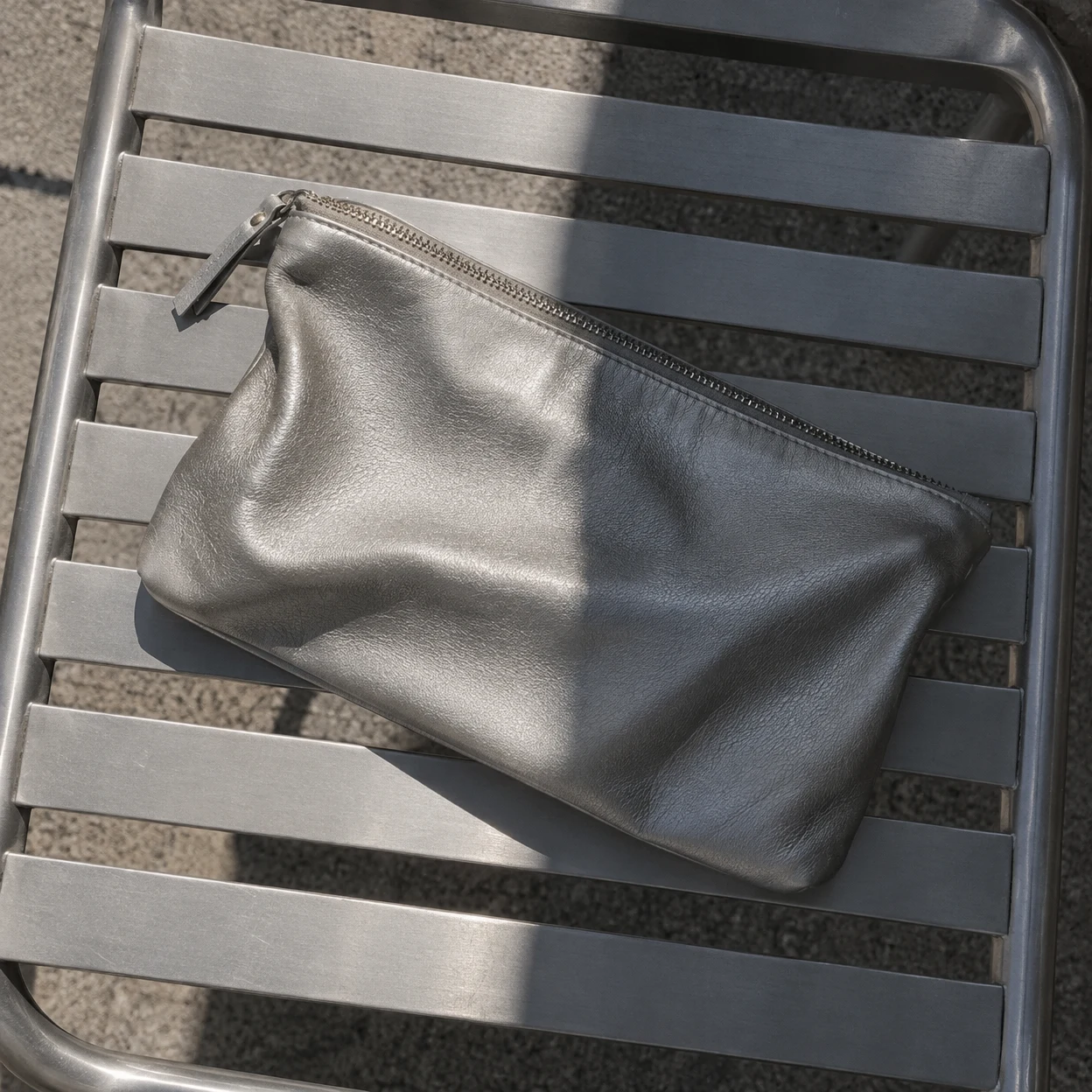 A softly folded silver leather zip clutch lies diagonally across the slatted seat of an aluminium chair, with sunlight and a sharp band of shadow crossing both…