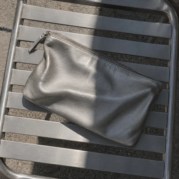 A softly folded silver leather zip clutch lies diagonally across the slatted seat of an aluminium chair, with sunlight and a sharp band of shadow crossing both…