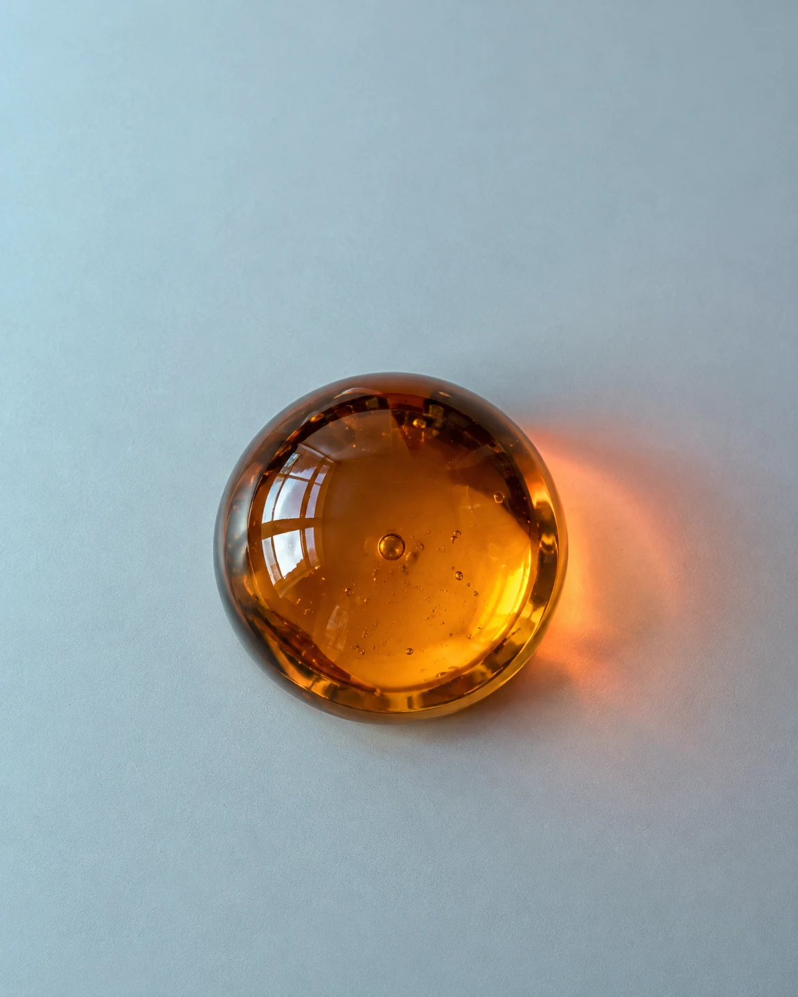 An amber hemispherical glass paperweight rests on a pale blue-grey tabletop, with a warm coral glow to its right and generous empty space above.