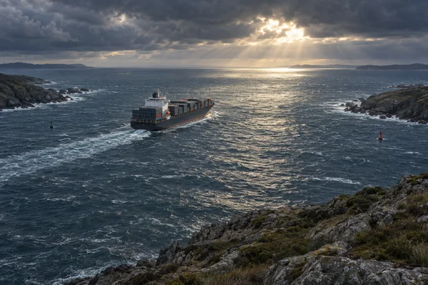 A container ship moves through choppy water between rocky coastal headlands beneath a break in heavy clouds, with sunlight reflecting across the sea and a…