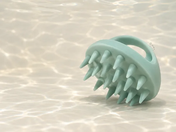 A seafoam silicone scalp brush sits at an angle on a pale ivory floor, its tapered teeth facing diagonally toward the camera and its grip arch visible behind…