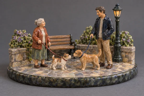 A glazed ceramic miniature plaza sits on a rounded paving base, with two full-length figures facing each other as their leashed dogs meet nose-to-nose; a…