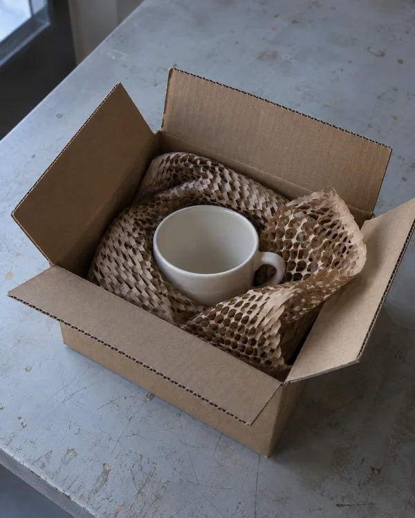 An open corrugated cardboard parcel sits diagonally on a worn pale-grey table, holding an ivory ceramic cup cushioned by expanded brown honeycomb paper.